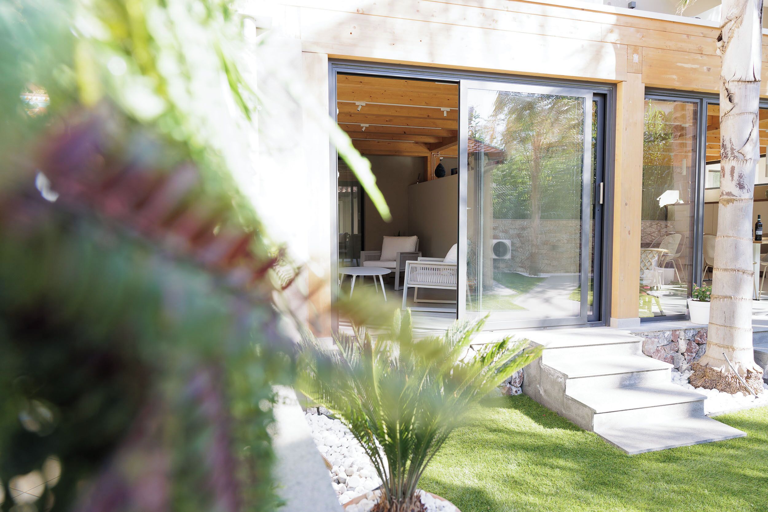 Une maison moderne dotée d’un patio, d’une porte vitrée donnant sur une pièce où se trouvent une chaise et une table blanches, ainsi que d’un petit jardin.