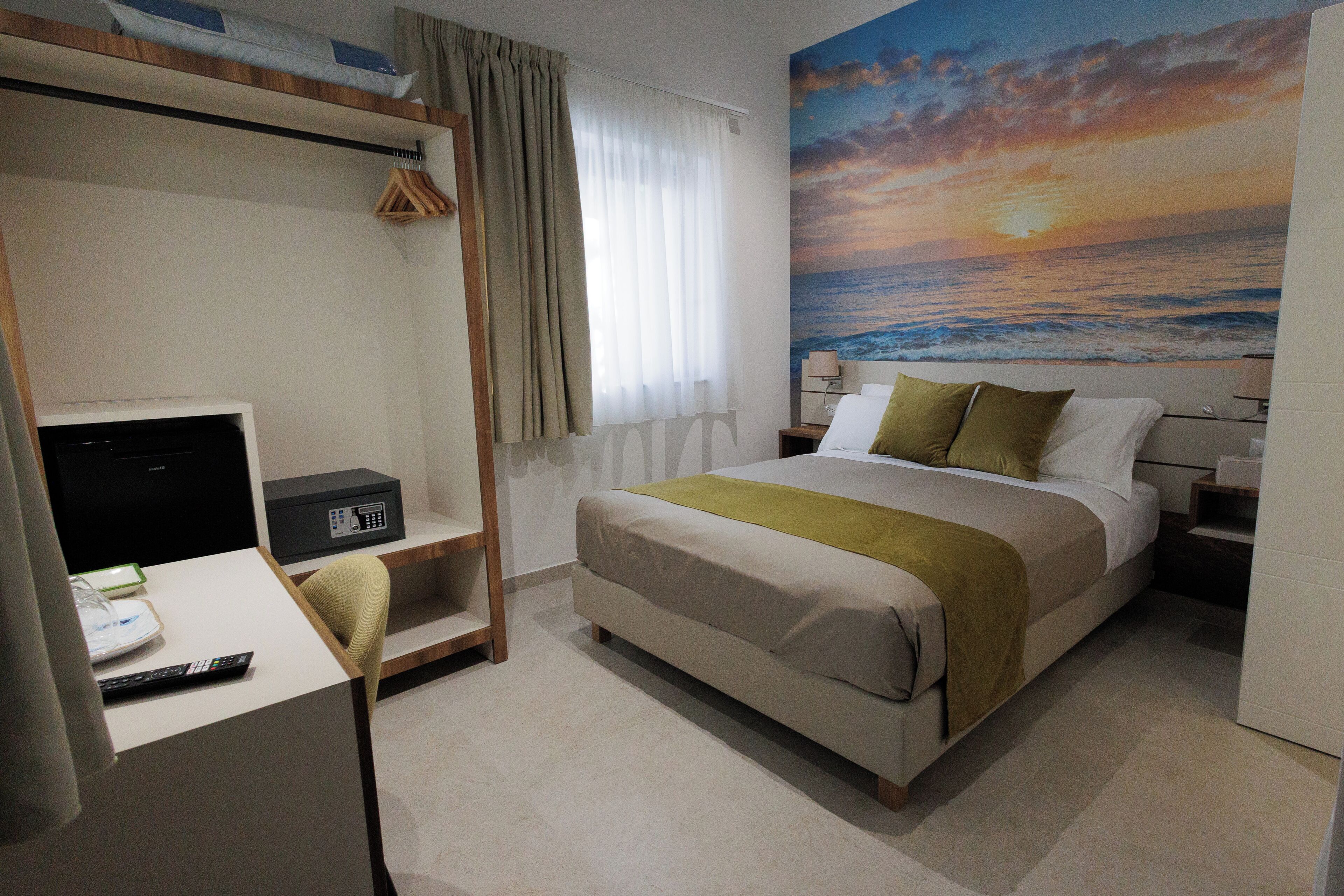 A hotel room with a bed, desk, chair, and a painting of a sunset over the ocean.