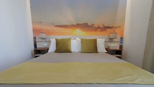 A hotel room with a bed, two nightstands, a lamp, and a sunset mural on the wall. - SEA GARDEN LUXURY ROOMS (Giardini Naxos)