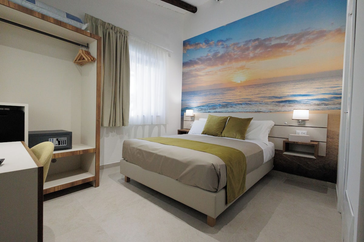 A hotel room with a bed, bedside tables, a desk, a chair, a wardrobe, and a large mural of a sunset over the ocean.