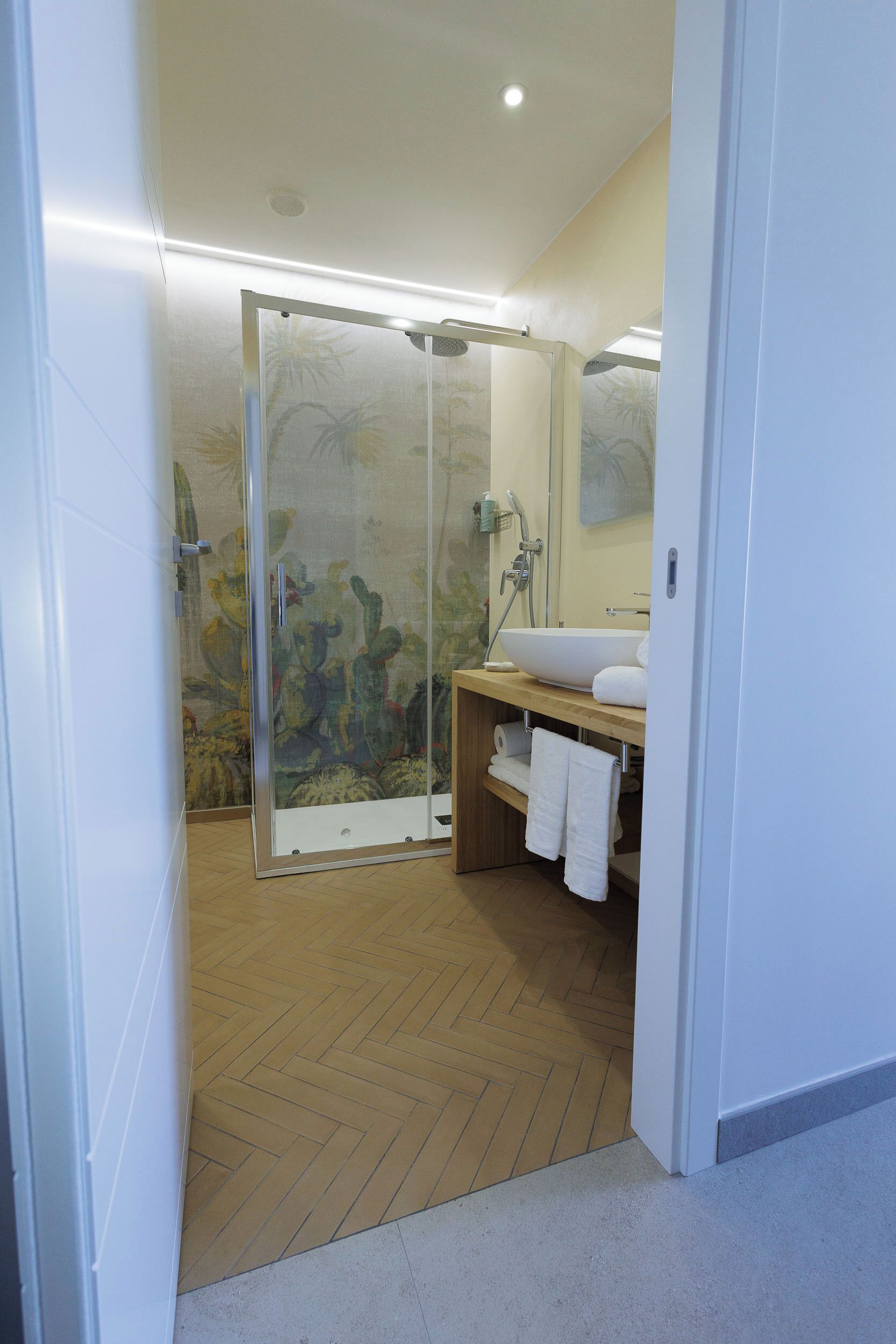 Een badkamer met een glazen douchecabine, een houten vloer, een witte wastafel en een spiegel.