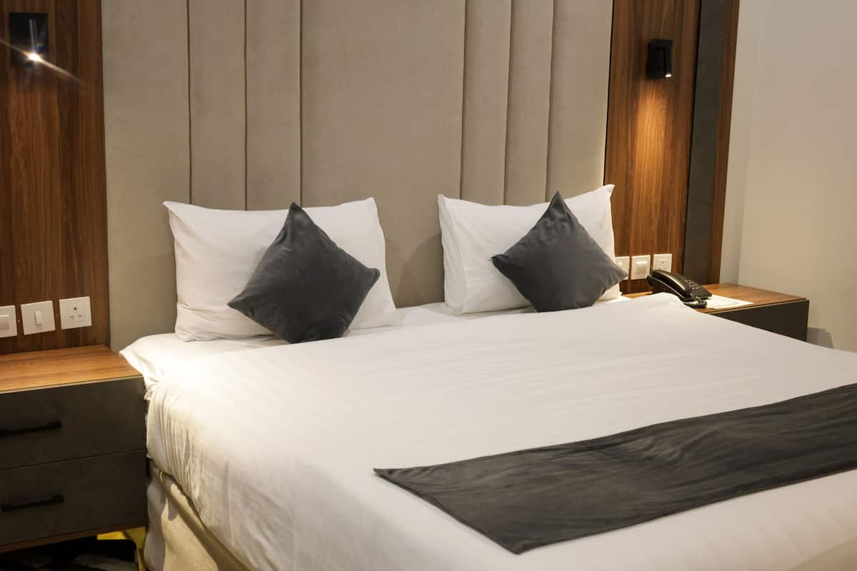 Una cama bien hecha con almohadas blancas y oscuras, un plaid oscuro y un cabecero de madera. Hay dos mesitas de noche, una con un teléfono y una lámpara, y la otra con un cajón.