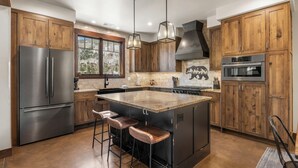 A kitchen with wooden cabinets, a stainless steel refrigerator, a large island with stools, pendant lighting, and a view through the window. - Lucky Star Lodge | Pool & Hot Tub | Fire Pit (Durango)