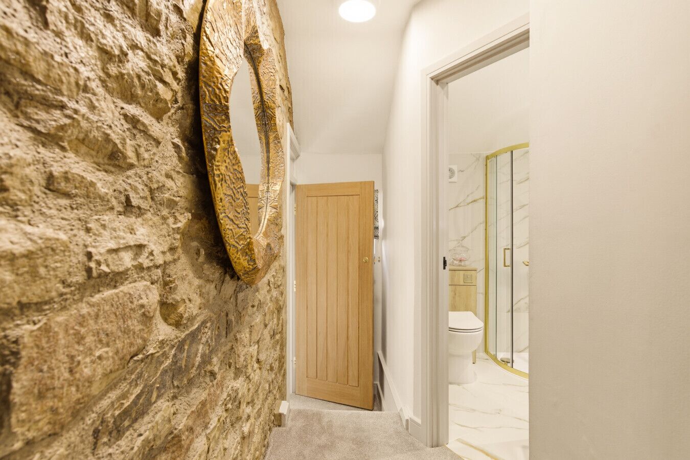 A hallway with a stone wall, a wooden door, and a bathroom with a marble floor and glass shower enclosure.