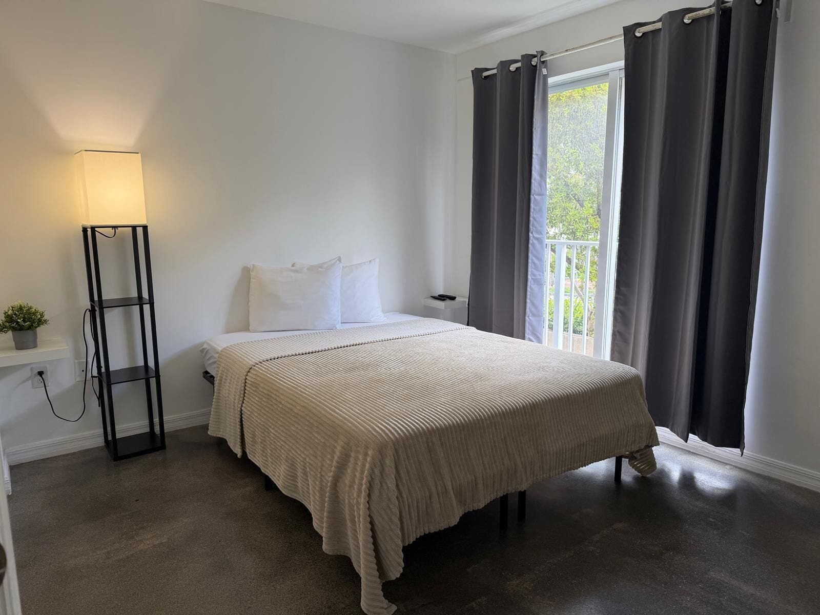 Un dormitorio con cama, mesita de noche, lámpara, ventana con cortinas y vista a zonas verdes.