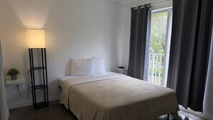 A bedroom with a bed, a bedside table, a lamp, a window with curtains, and a view of greenery outside. - Triangle Suites LLC (Hialeah)