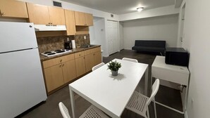 A compact kitchen with a refrigerator, stove, and wooden cabinets. There is a dining table with chairs and a small potted plant. A black microwave is on a counter, and a dark sofa is in the background. - Triangle Suites LLC (Hialeah)