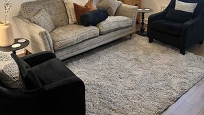 Ein Wohnzimmer mit gepolstertem Sofa mit Muster, einem schwarzen Sessel, einem runden Spiegel, einer Hängeleuchte und einem Fenster mit Jalousien.