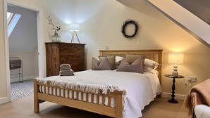 Ein Schlafzimmer mit einem hölzernen Bett, einem Schminktisch, einem kleinen Tisch mit Lampe, einem Korbstuhl und Blick auf einen Balkon.