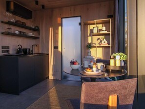 A modern kitchen with a dark countertop, wooden cabinets, and a glass door leading to a dining area with a round table, chairs, and a small shelf with decorative items.