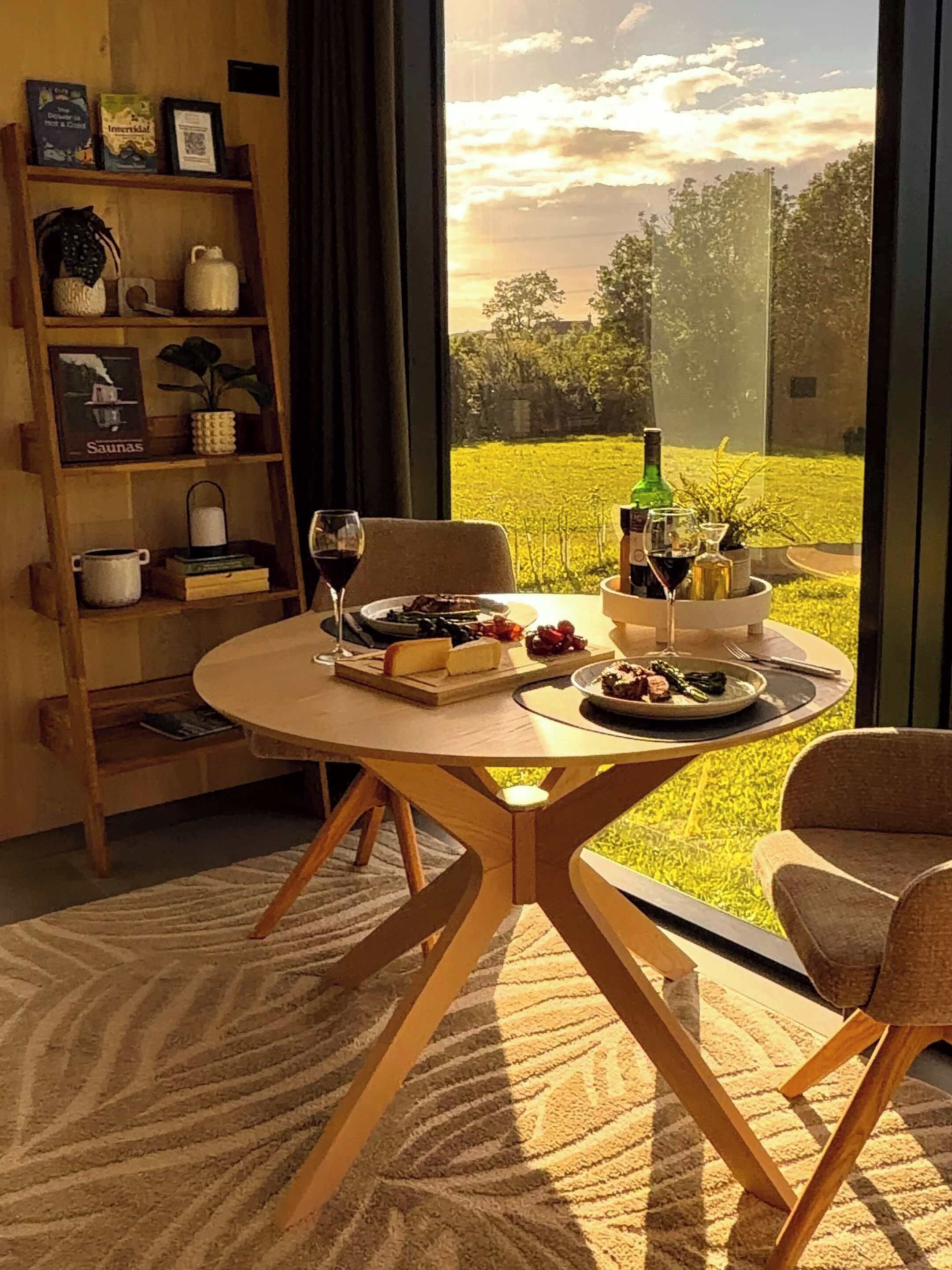 A dining table with a view of a field, a bookshelf with books and decorative items, and a window with curtains.