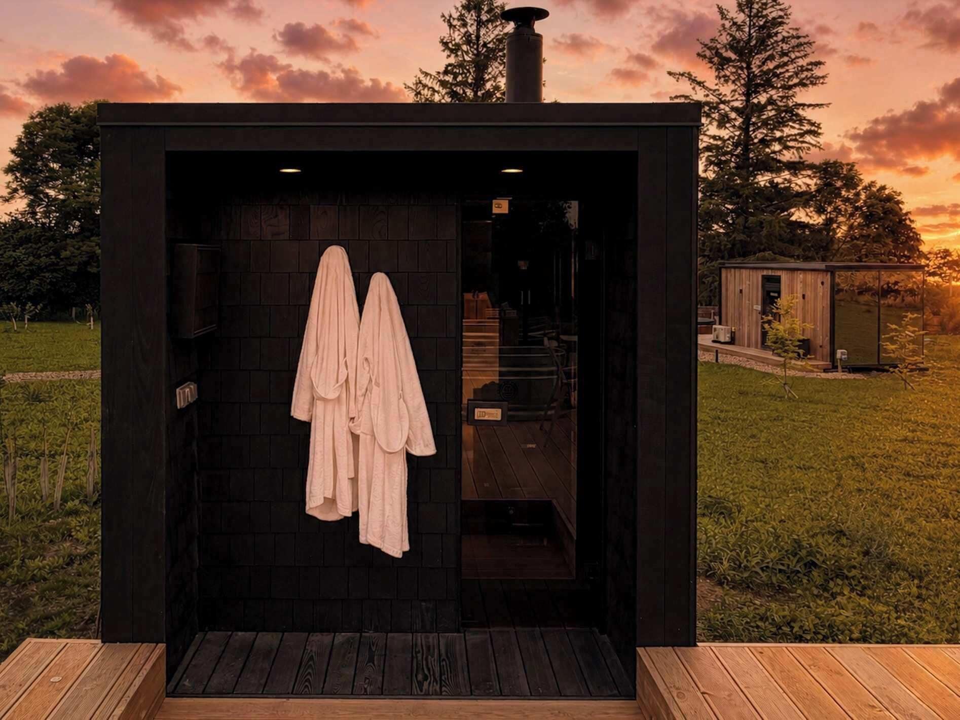 A modern, dark-colored sauna with a wooden deck entrance, a visible towel rack, and a small adjacent structure.