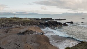 Paisaje costero con terreno rocoso, un faro y montañas al fondo.