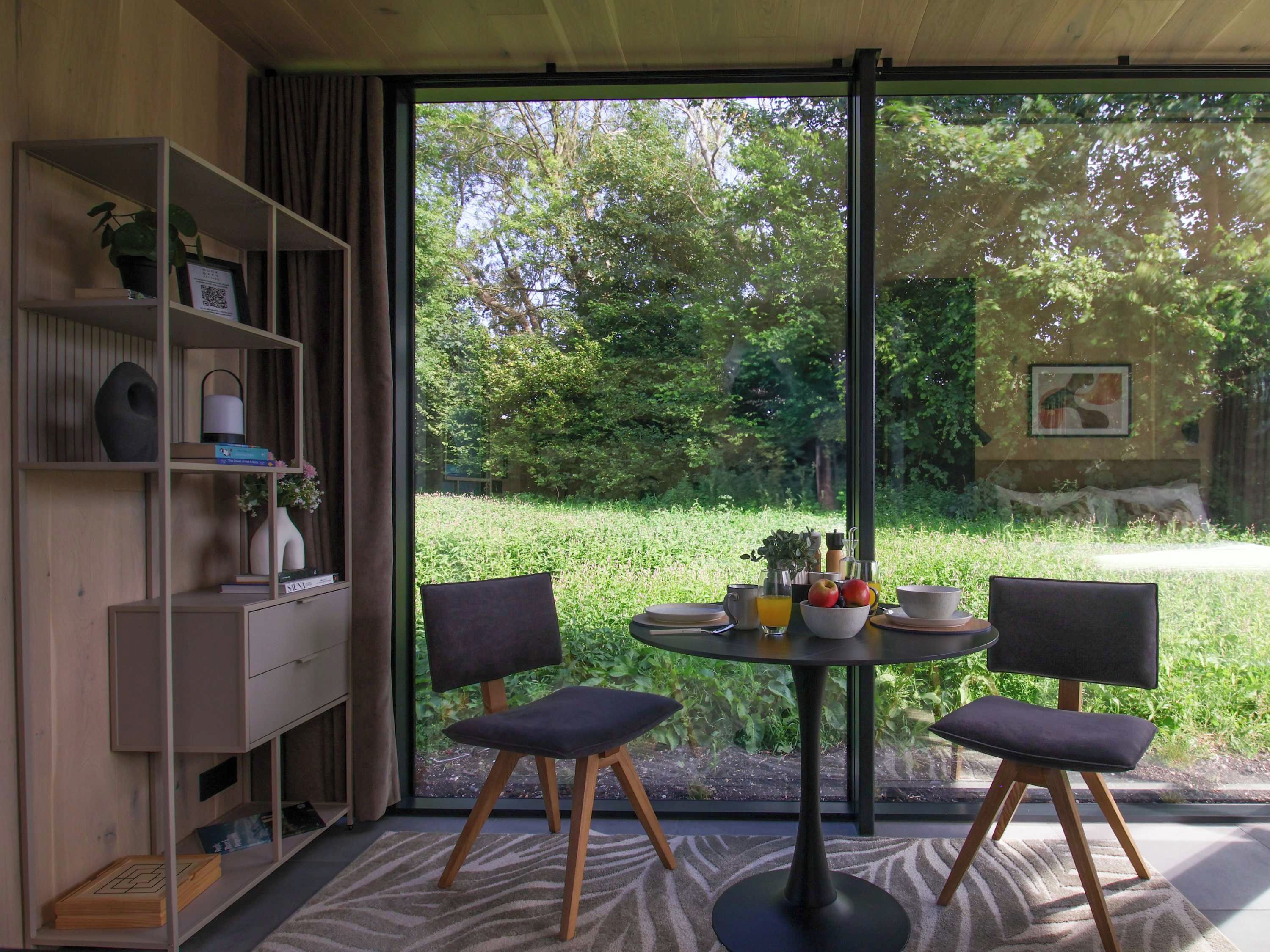 A room with a large window, a round table with chairs, a bookshelf, and a painting on the wall.