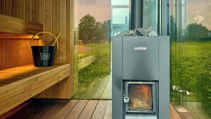Una sauna con una stufa a legna Harvia, una panca in legno e una parete di vetro che offre vista su un paesaggio verde.
