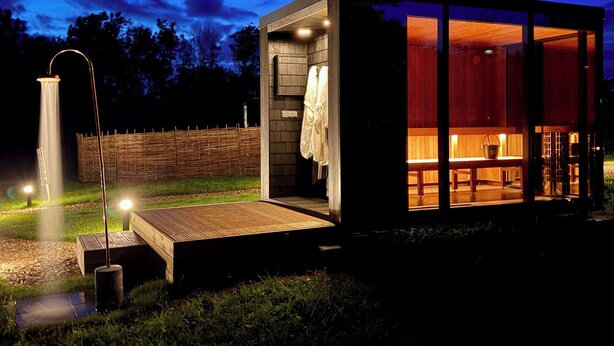 Een moderne, goed verlichte buitendouche met een houten terras, een sauna met glazen wanden, een bankje en een emmer, en een houten hek op de achtergrond.