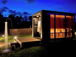 A modern, well-lit outdoor shower area with a wooden deck, a glass-enclosed sauna room with a bench and a bucket, and a wooden fence in the background.
