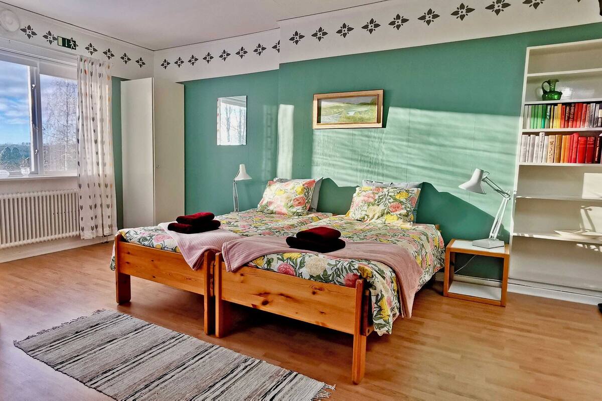 Ein Schlafzimmer mit einem Holzbett, einem Schreibtisch, einem Stuhl, einem Bücherregal und Blick ins Freie.