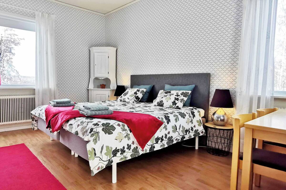 Ein Schlafzimmer mit Bett, Nachttisch, Stuhl, Tisch, Heizkörper und einem Fenster mit Vorhängen.
