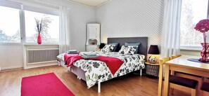 Une chambre à coucher comprenant un lit, une table de chevet, une chaise, une table, un radiateur et une fenêtre avec des rideaux.