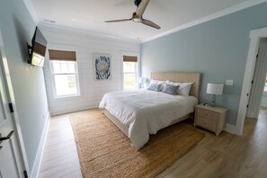 A bedroom with a large bed, a nightstand, a ceiling fan, and a wall-mounted TV. - Happy Tides Hideaway/NEW!/Pool/Kids Adventure Room (Oak Island)