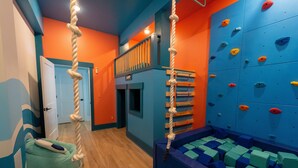 A climbing gym with a rope climbing wall, padded flooring, and a padded seating area. - Happy Tides Hideaway/NEW!/Pool/Kids Adventure Room (Oak Island)