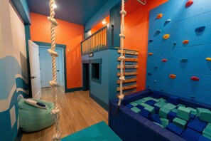 A climbing gym with a rope climbing wall, padded flooring, and a padded seating area. - Happy Tides Hideaway/NEW!/Pool/Kids Adventure Room (Oak Island)