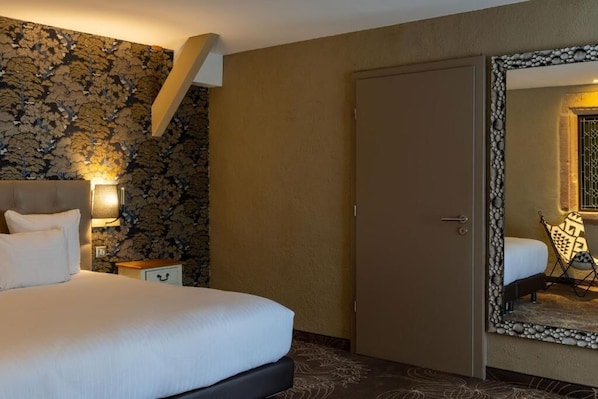 A hotel room with a bed, a nightstand, a wall with a floral pattern, a door, and a mirror with a decorative frame. - Hotel Restaurant l'Orchidée (Ensisheim)