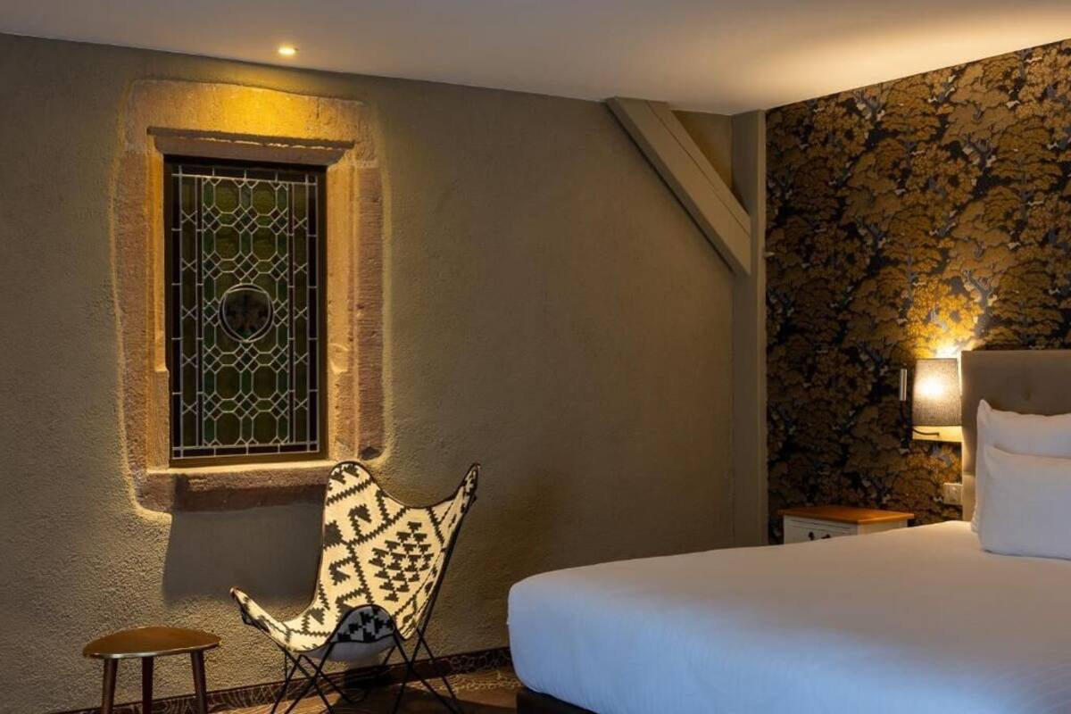 Une chambre à coucher comprenant un lit, une chaise, une petite table, une fenêtre ornée d’une grille décorative et un mur recouvert d’un papier peint à motifs.