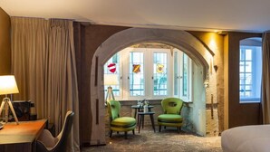 A hotel room with a bed, a desk with a lamp, two chairs, a small table with a vase, and a view through an arched doorway. - Hotel Restaurant l'Orchidée (Ensisheim)