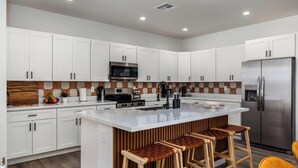 Uma cozinha moderna com armários brancos, uma geladeira de aço inoxidável, um micro-ondas e uma bancada grande com banquetas.
