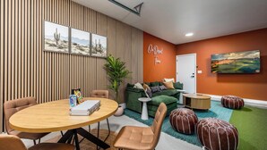 Quarto moderno com mesa de madeira redonda, duas cadeiras, sofá verde, mesinha lateral branca e planta verde.
