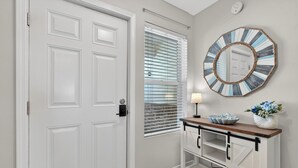 Une porte blanche, une fenêtre avec des stores, un miroir fixé au mur, une console en bois avec une lampe et un vase rempli de fleurs.