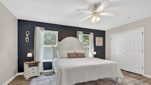 Une chambre avec un lit blanc, un ventilateur de plafond, une fenêtre avec des stores, une porte et une table de chevet avec une lampe.