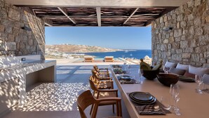 A dining area with a view of the sea, stone walls, and a long table set for dining. - Sky Resort in Mykonos (Mykonos)
