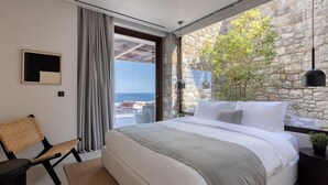 A modern bedroom with a large bed, a view of the sea, a stone wall, and a hanging light fixture. - Sky Resort in Mykonos (Mykonos)