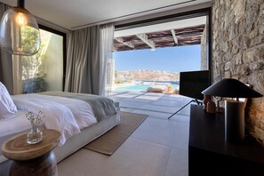 A bedroom with a bed, a bedside table, a lamp, and a view of a pool and landscape. - Villa Sky II in Mykonos (Mykonos)