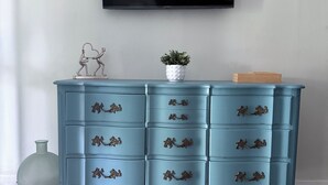 Uma cômoda azul-turquesa com várias gavetas, um vaso de planta e um objeto decorativo de metal.