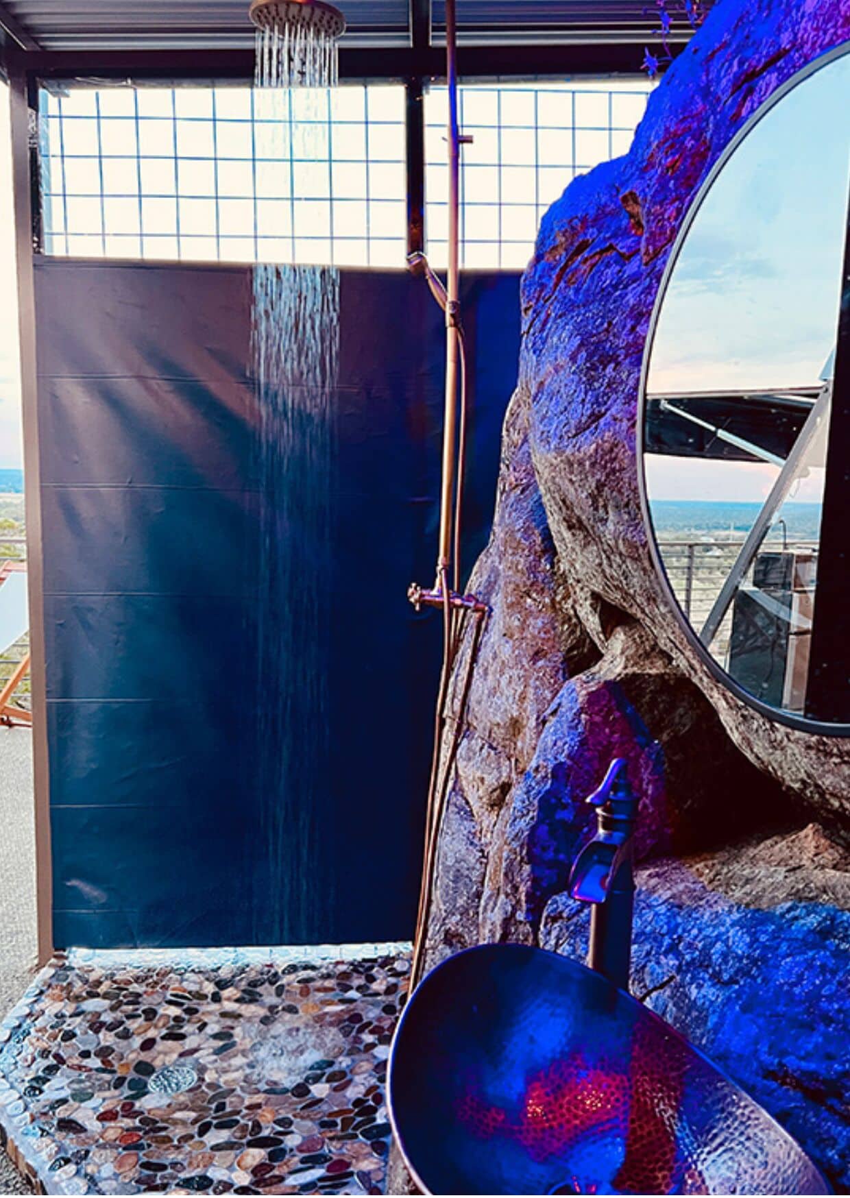 A bathroom with a stone mosaic floor, a large mirror, and a shower with a waterfall feature.