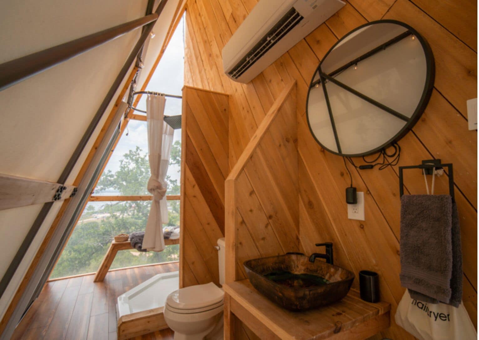 Une cabane en bois avec un toit en pente, des toilettes, un lavabo avec un receveur, un appareil de climatisation et une serviette suspendue à un porte-serviettes.