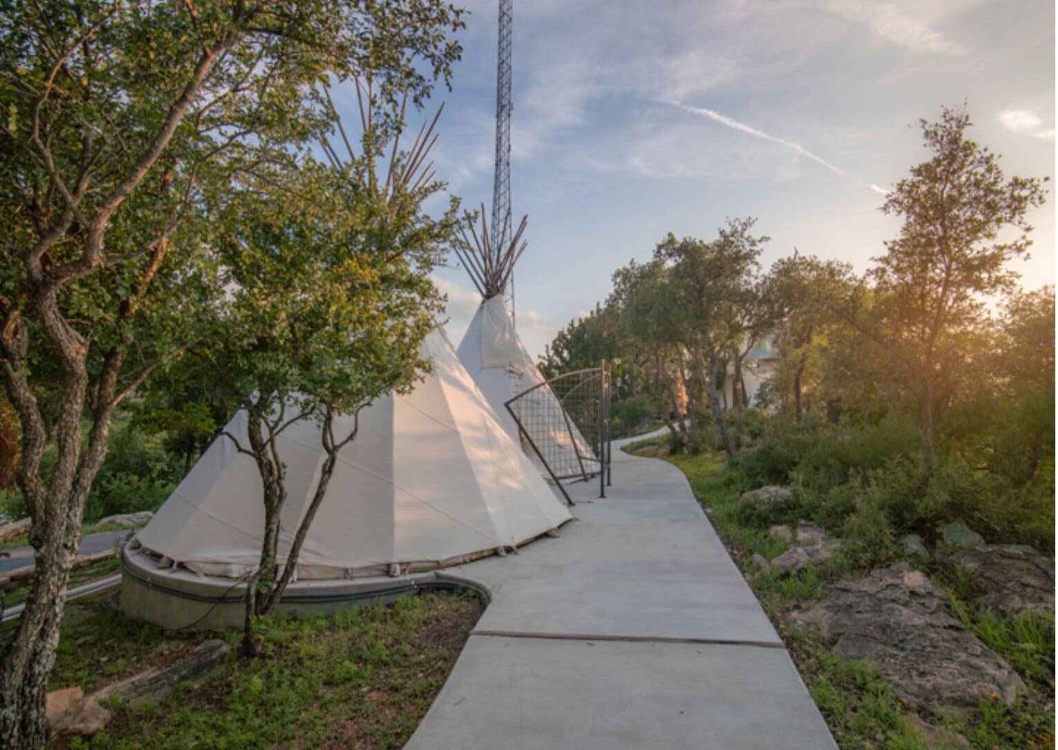 A white tent-like structure with a metal staircase leading up to it, surrounded by trees and a concrete pathway.