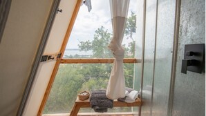 Una stanza con pavimento in legno, un tavolino con un cesto e una coperta, e vista su alberi e acqua attraverso una finestra con tenda.