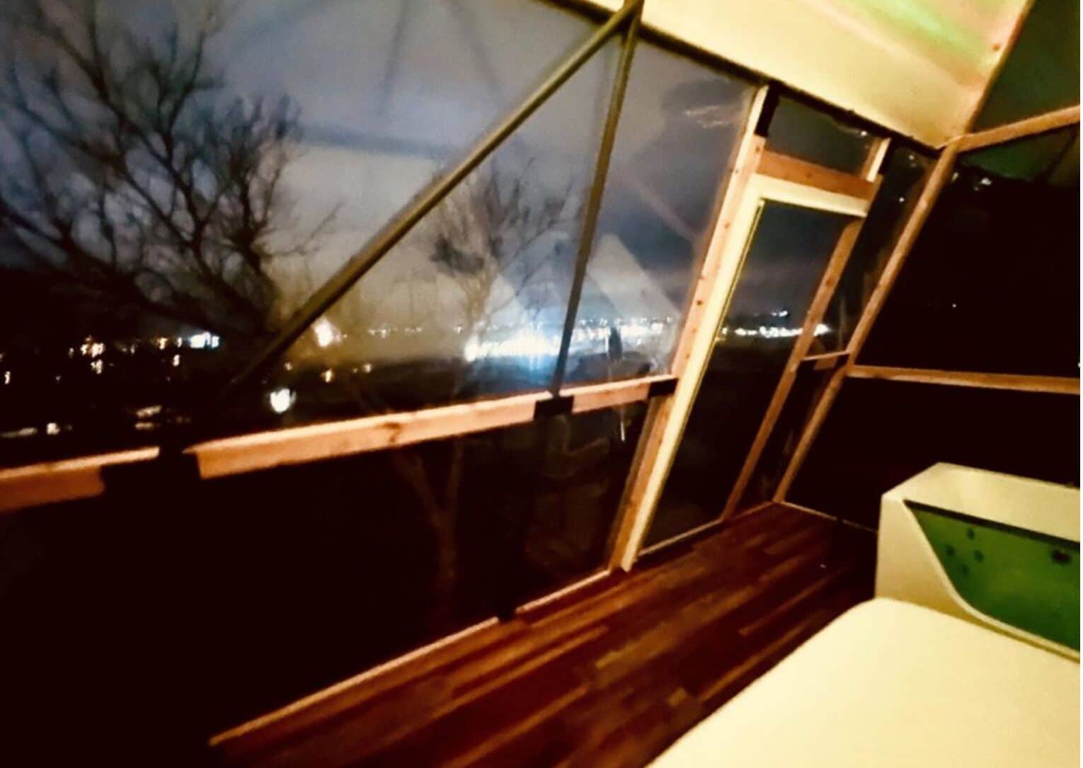A room with a view of a cityscape at night, a window with a wooden frame, and a white surface with a green screen.