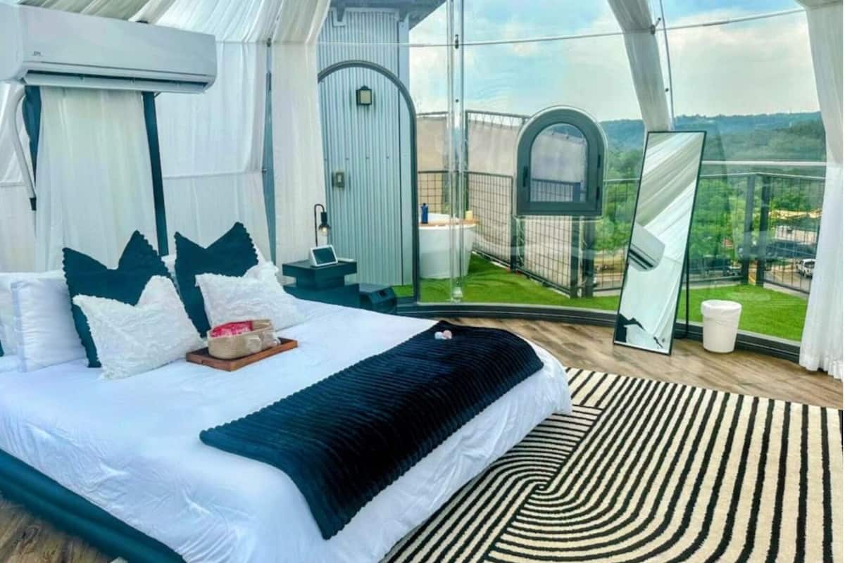 Une chambre à coucher avec un lit, des oreillers, une table de chevet, un miroir et une vue sur l’extérieur.