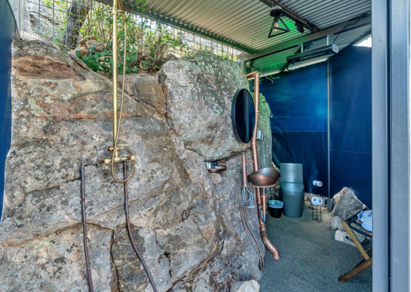 A rustic outdoor kitchen with stone walls, copper pipes, and a hanging pot rack.