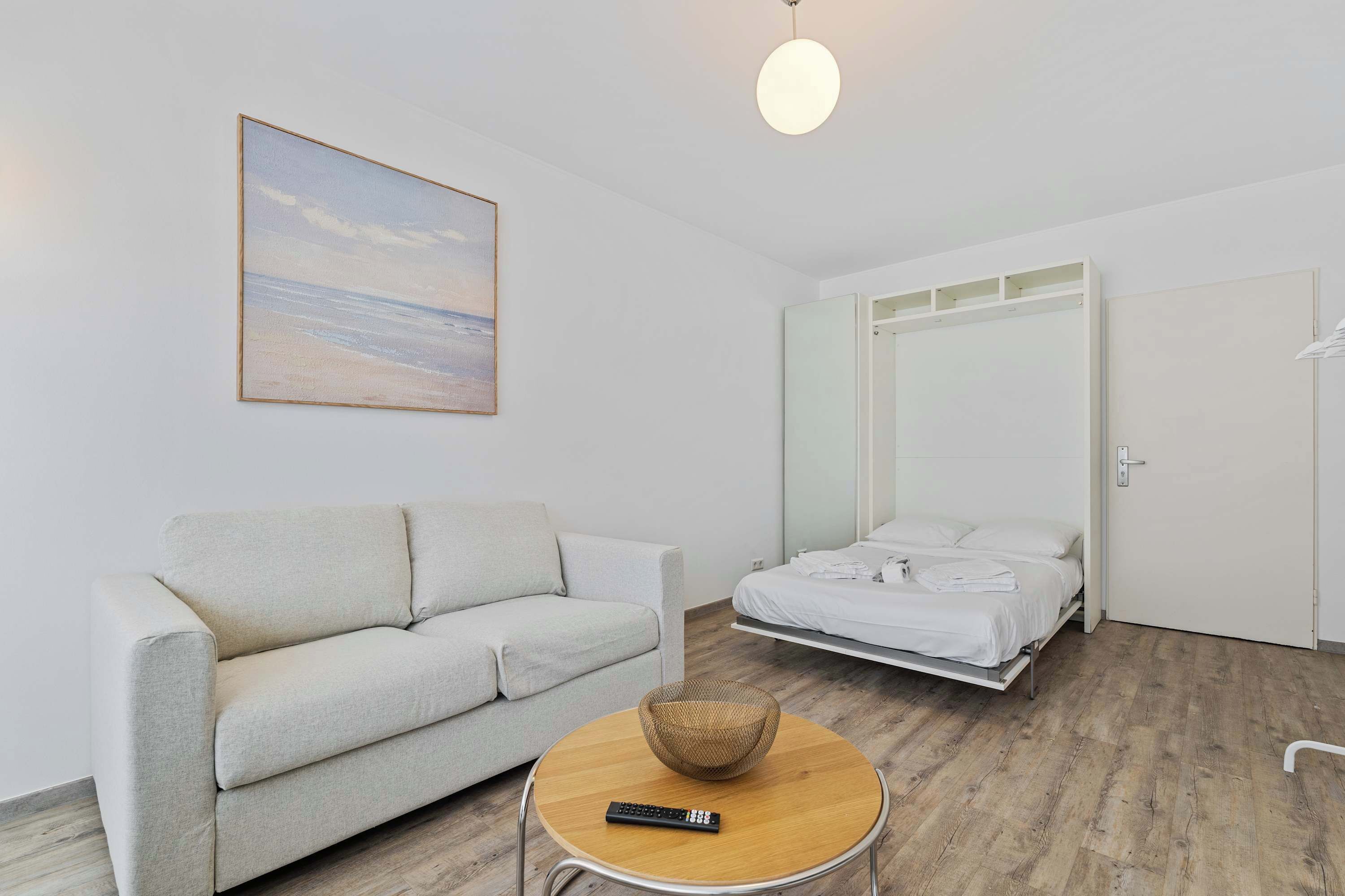 Una habitación moderna con sofá, cama con cabecera, una mesita redonda con una cesta y un cuadro en la pared.