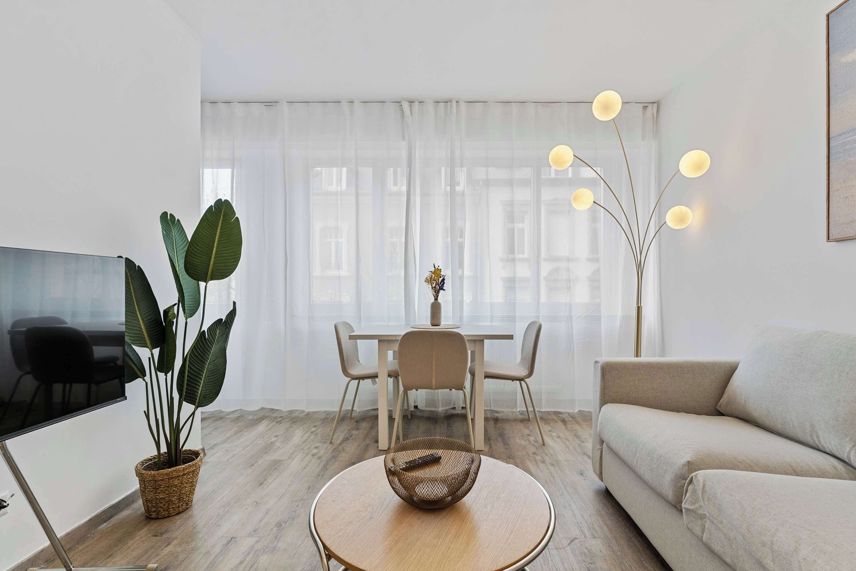 Un salon moderne comprenant un canapé, un téléviseur, une table à manger avec des chaises, une grande plante et un lampadaire original.
