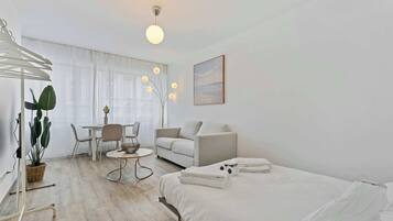 Una habitación de hotel moderna con una cama, un sofá, una mesa de comedor con sillas, una mesita con un jarrón y un gran ventanal con cortinas.