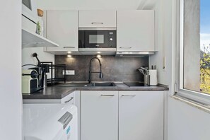 A modern kitchen with white cabinets, a built-in microwave, a stainless steel sink, and a window with a view of trees. - Modern Studio in Central Luxembourg City (Luxembourg)