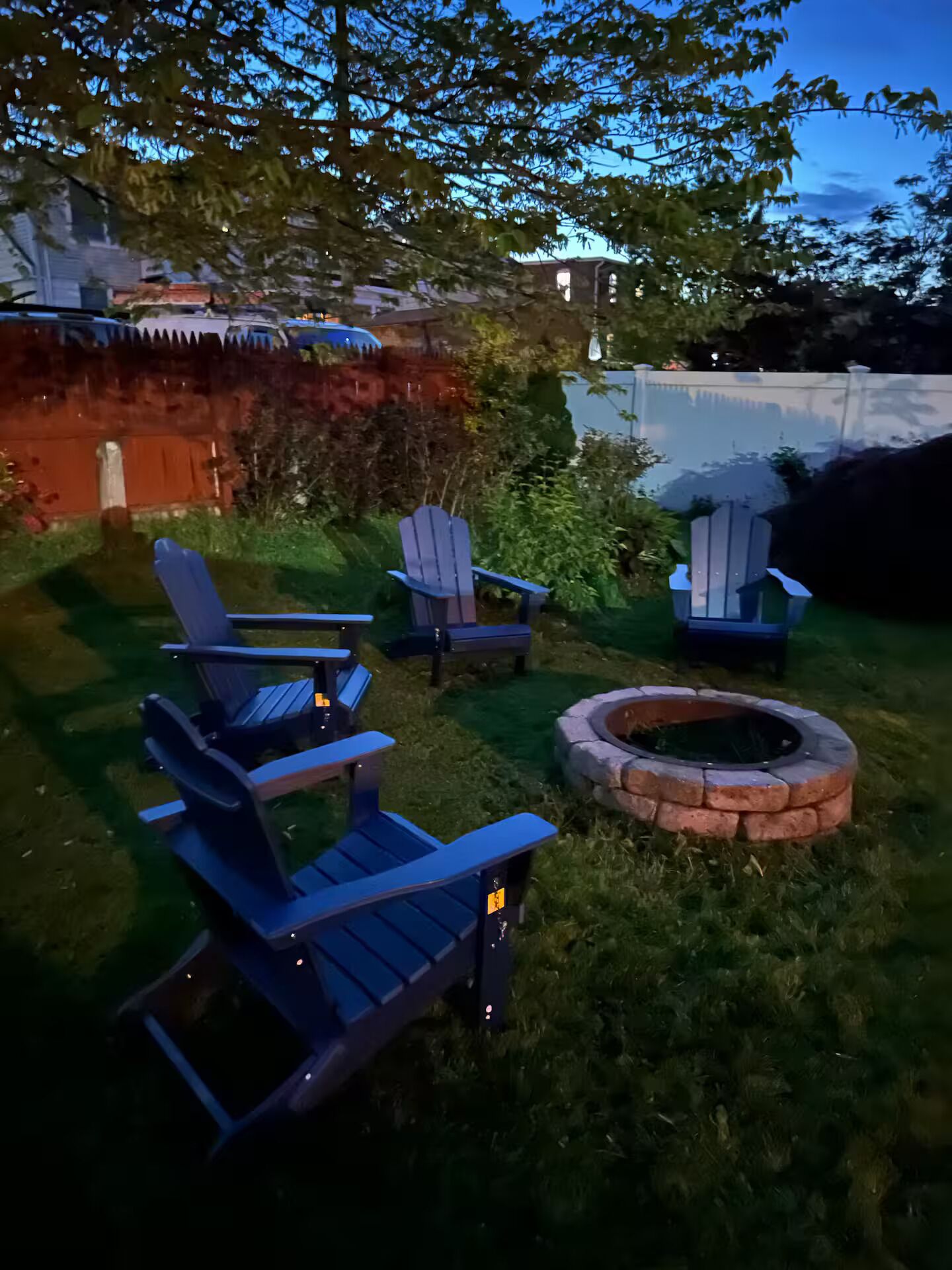 A backyard with four Adirondack chairs, a stone fire pit, and a red fence.